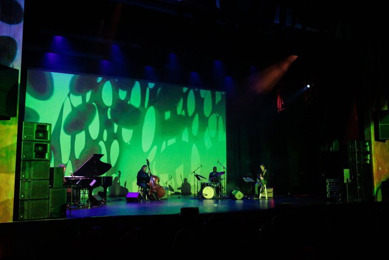 A jazz trio performs on a dark stage before a large screen displaying abstract green shapes.