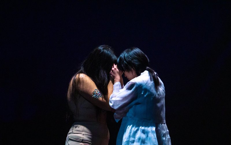 Two figures in dim light hold hands against a dark background. One wears a light top and pants, with a jeweled arm piece. The other wears a white, long-sleeved dress.