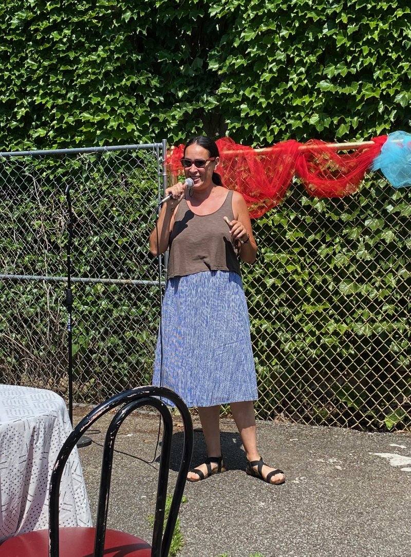 Woman in sunglasses, brown top, and blue striped skirt, holding a microphone, stands before a chain-link fence with red and blue netting.