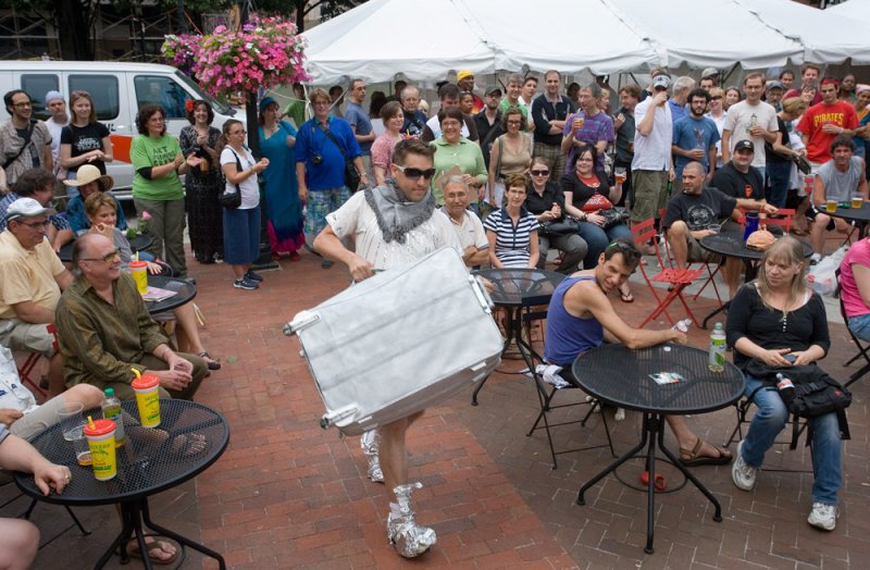 Man in sunglasses and tin foil boots carries a large silver suitcase through an outdoor art event.