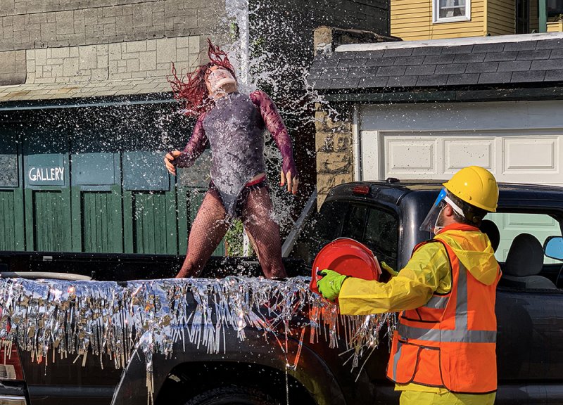 A person in a dark bodysuit and red wig is doused with water from a red bucket by another person in a yellow suit.