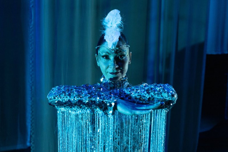 Woman in an elaborate, shimmering blue costume with a feathered headpiece, looking forward with blue lighting.