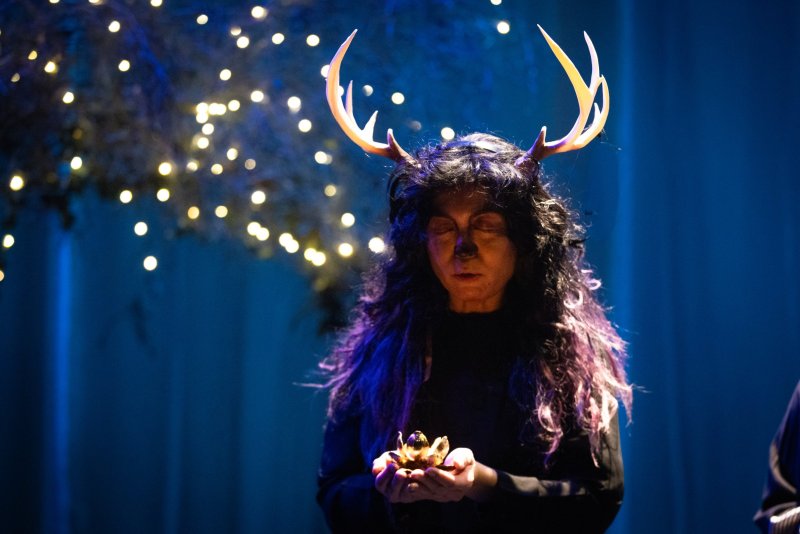 Person with antlers and long dark hair, eyes closed, holding a small golden object, against a blue background with warm string lights.