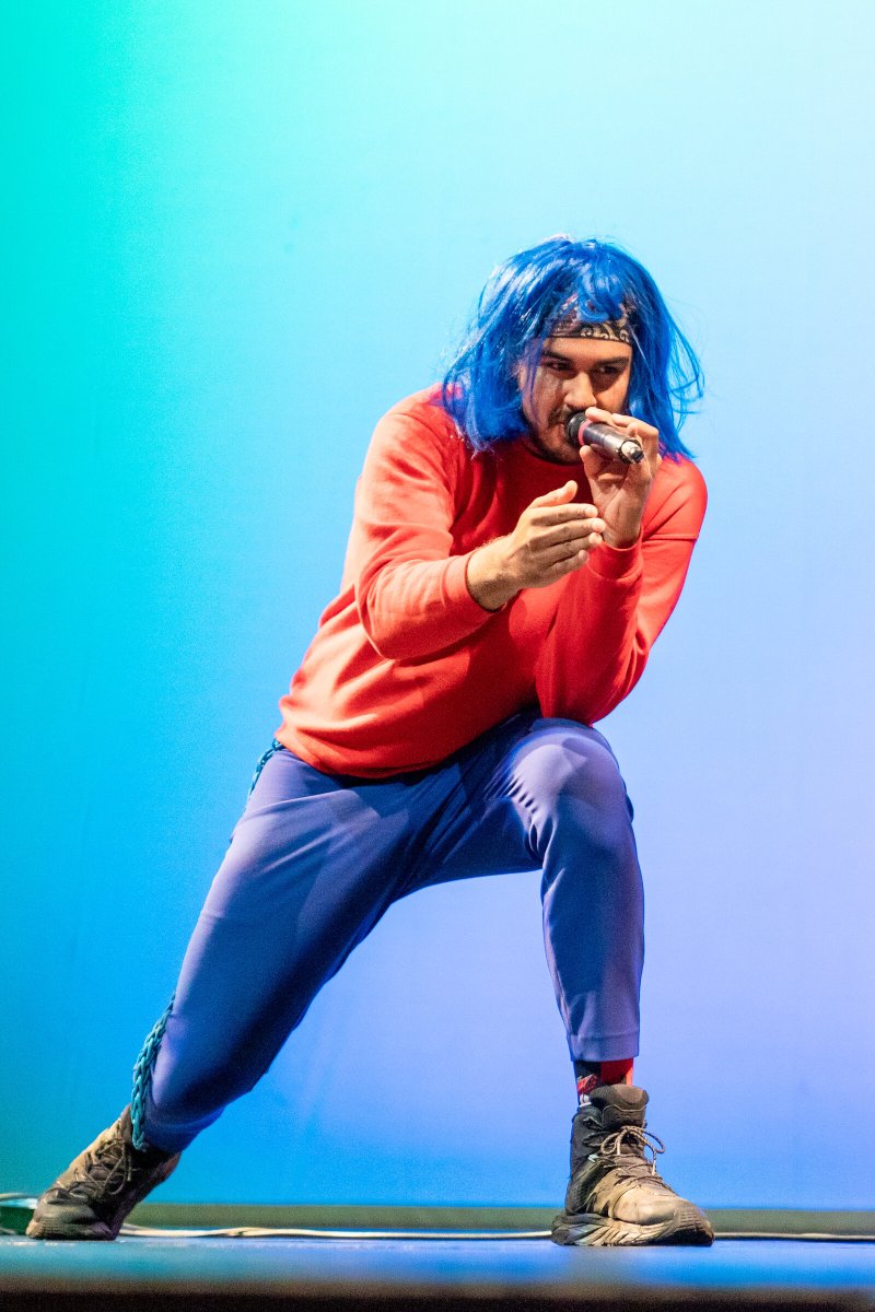 Person in blue wig, red shirt, and blue pants kneels, singing into a microphone against a blue-green gradient background.
