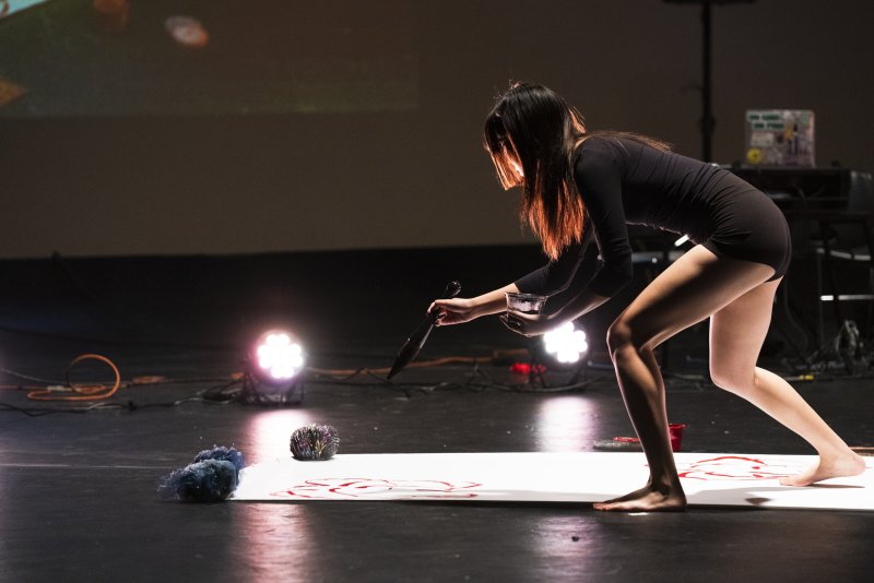 A barefoot woman in a black leotard paints red designs on a white canvas on a dark stage with bright lights.