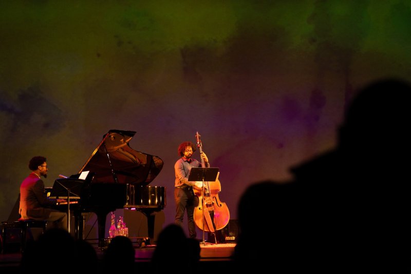 Two musicians, one on grand piano and one on upright bass, perform on a stage with a mottled green and purple backdrop.