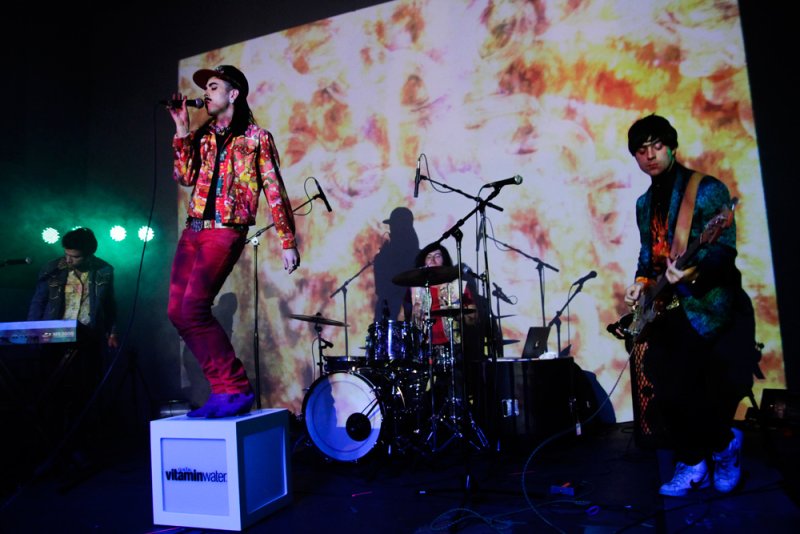 Band performing on stage with colorful backdrop. Singer stands on a Vitaminwater box.