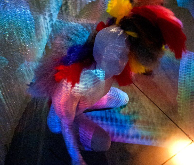 Sculptural figure made of translucent mesh, illuminated by colorful lights, with red, yellow, and blue accents.