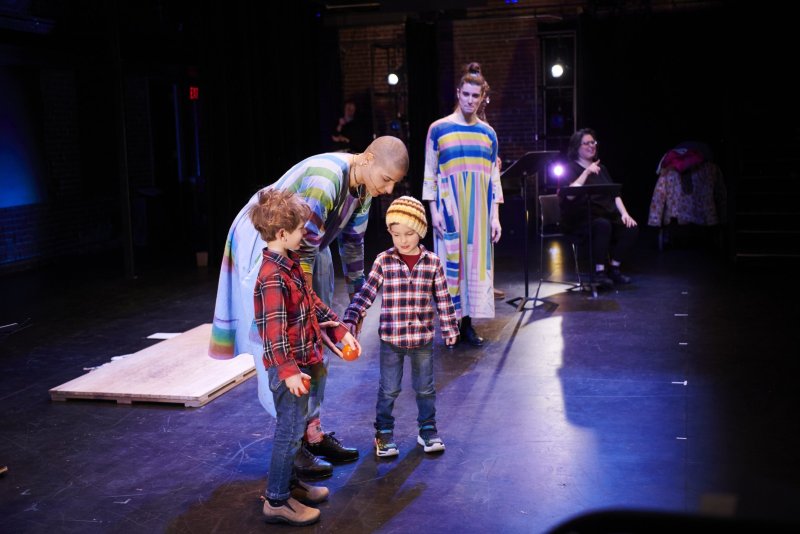 A stage scene with two children in plaid shirts holding orange spheres, flanked by adults in colorful striped dresses. A woman sits in the background. The stage floor is dark, lit with blue and purple hues.