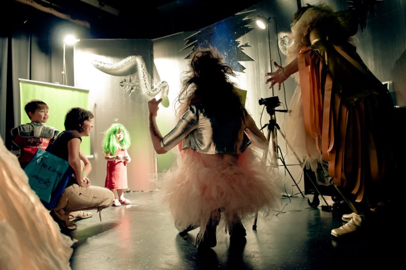 Performance art with a person in a pink tutu and silver top facing a green-haired child, while two people watch.
