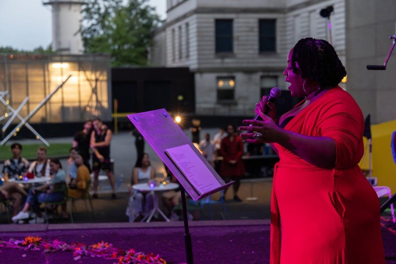 Outdoor performance scene: a Black woman in a red dress sings into a microphone. A purple-lit music stand is nearby. In the background, people sit at tables, with a building visible behind them.