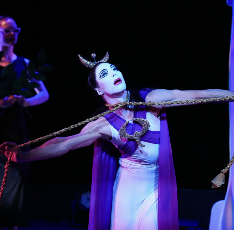 Performer in white and purple costume, elaborate makeup, and horned headdress, pulling a golden rope.