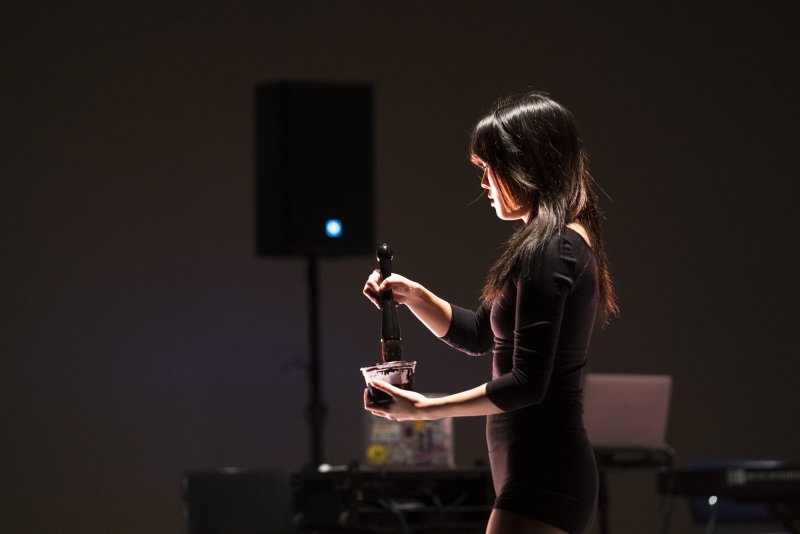 A woman in profile dips a large brush into a container. She is illuminated by a spotlight. Behind her are a speaker, a laptop, and a keyboard, all out of focus. The background is dark.