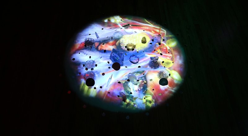 Projected circular light with scattered objects, including clear and dark stones, against a colorful, abstract background.
