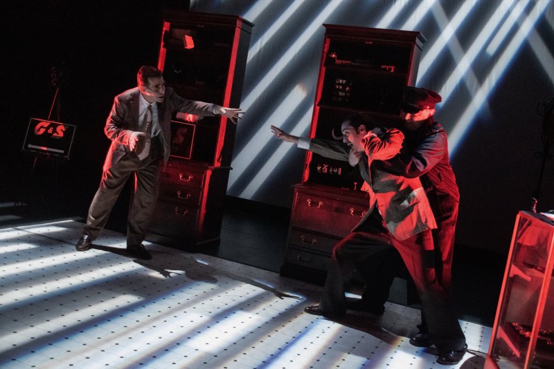 Three men in a dramatic scene with red lighting. One man in a suit gestures, another is held by a third in a cap.