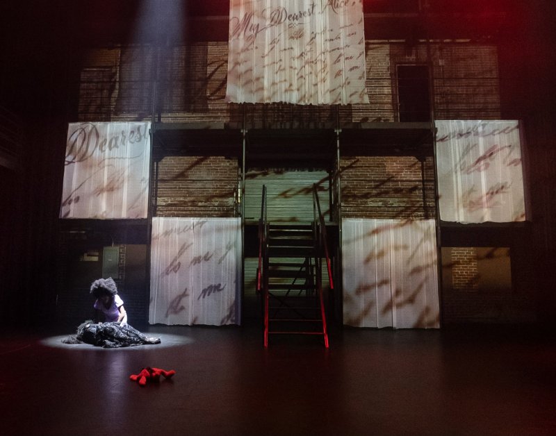 Stage set with brick wall backdrop, hanging translucent panels with letter projections, red stairs, and figure in spotlight.
