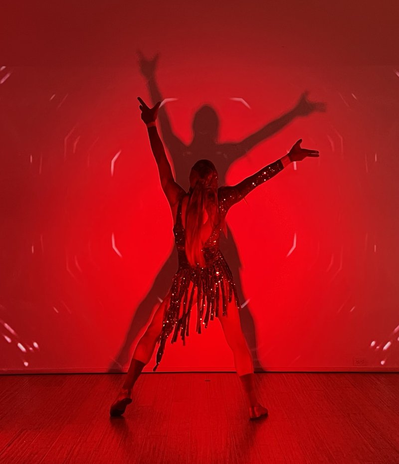 Dancer in a sparkling costume with arms outstretched, silhouetted against a vibrant red background with a faint, larger shadow behind.