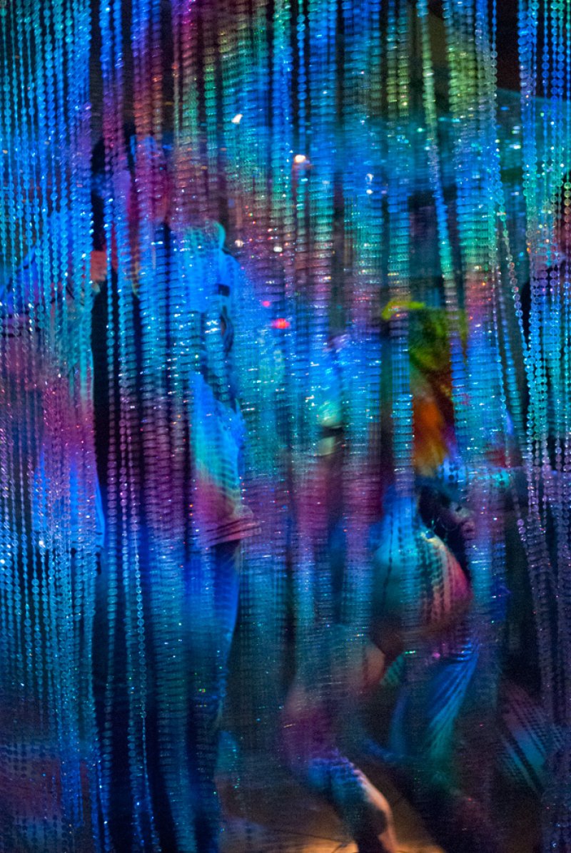 Vertical strands of blue, green, and pink beads create a shimmering, ethereal curtain, with blurred figures behind.