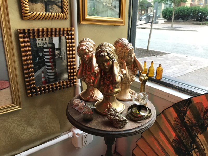 Three golden busts of women with closed eyes on a round table, surrounded by ornate frames and a street view.