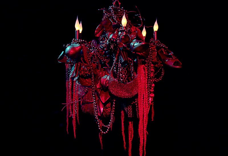 Sculptural assemblage in red light against black. Features shoes, beads, foliage, and lit candles in a dense, ornate composition.