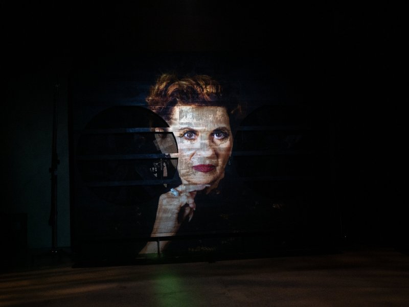 Projection of a woman with red lips and brown hair, chin resting on her hand, on a dark, ribbed surface.