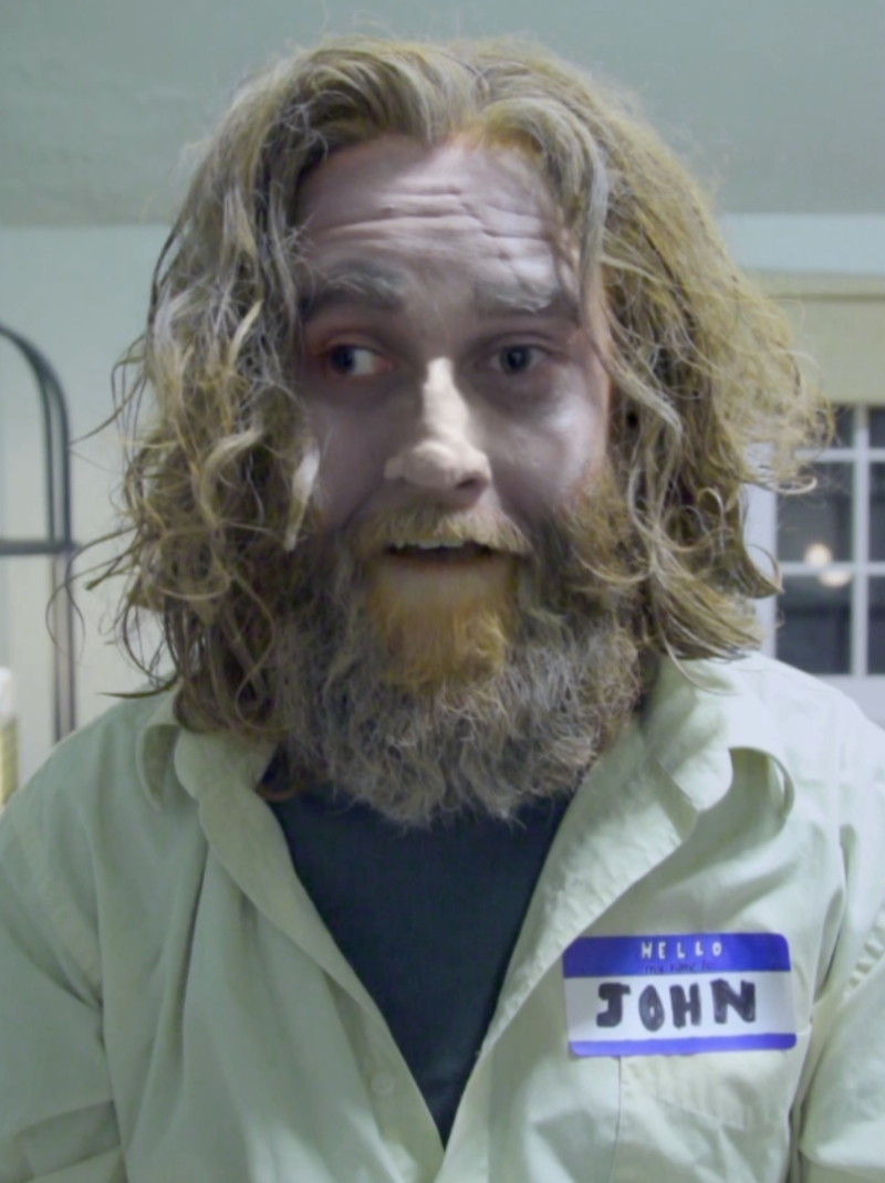 Man with long, messy brown hair and beard, pale face, wearing a light shirt and "Hello John" name tag.