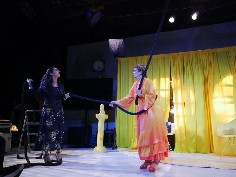 Two women on a stage, pulling a thick black rope. One wears a dark floral dress, the other a flowing orange gown.