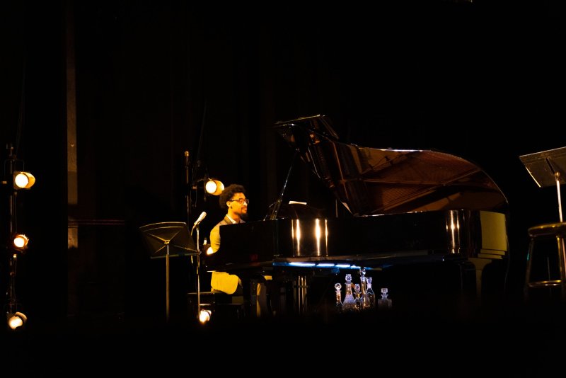 A man with glasses and curly hair plays a grand piano on a dark stage, illuminated by warm spotlights.