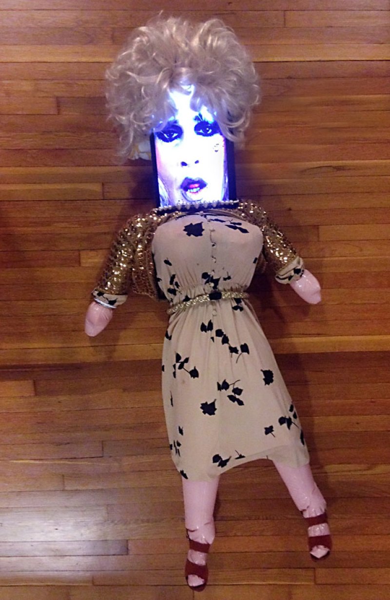Doll with a screen for a head displaying a drag queen's face, blonde wig, gold sequin jacket, and floral dress.