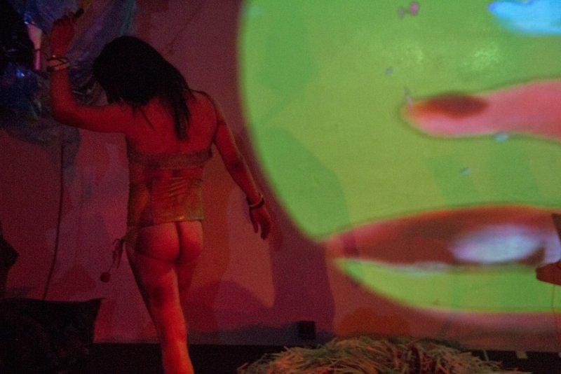 Woman in gold corset and thong, seen from behind, dancing in front of a green and red projected light.