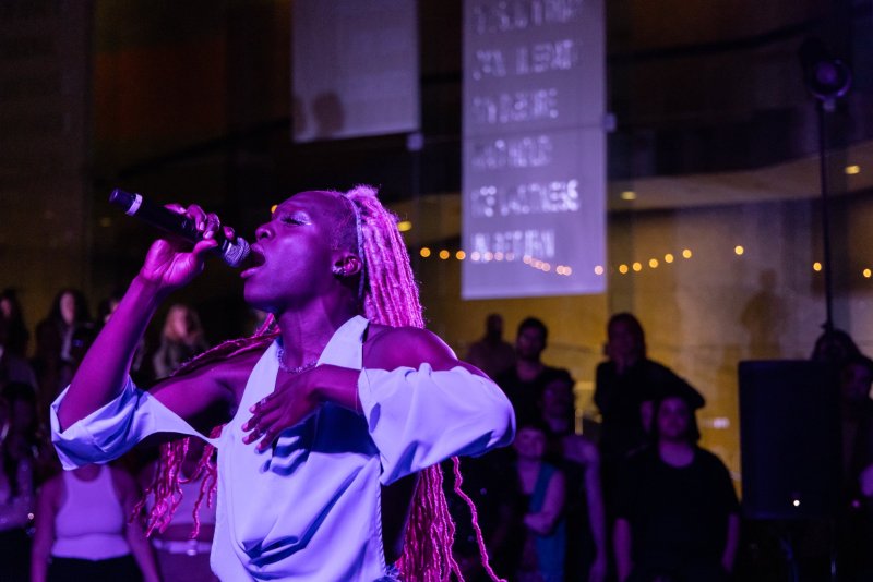 A Black performer with pink dreadlocks sings into a microphone on stage, bathed in purple light, with text projected on banners in the background.