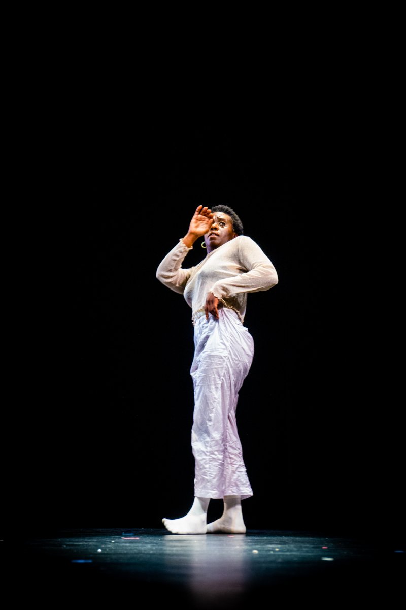 A person in white clothing stands on stage with a black background, shielding their eyes.