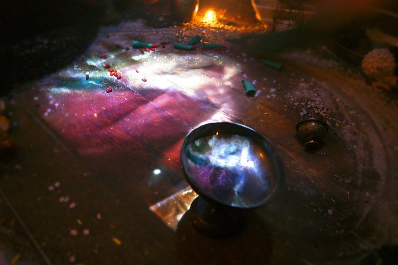 A dark, high-angle shot of a séance table with a reflective bowl, scattered objects, and colorful light projections.