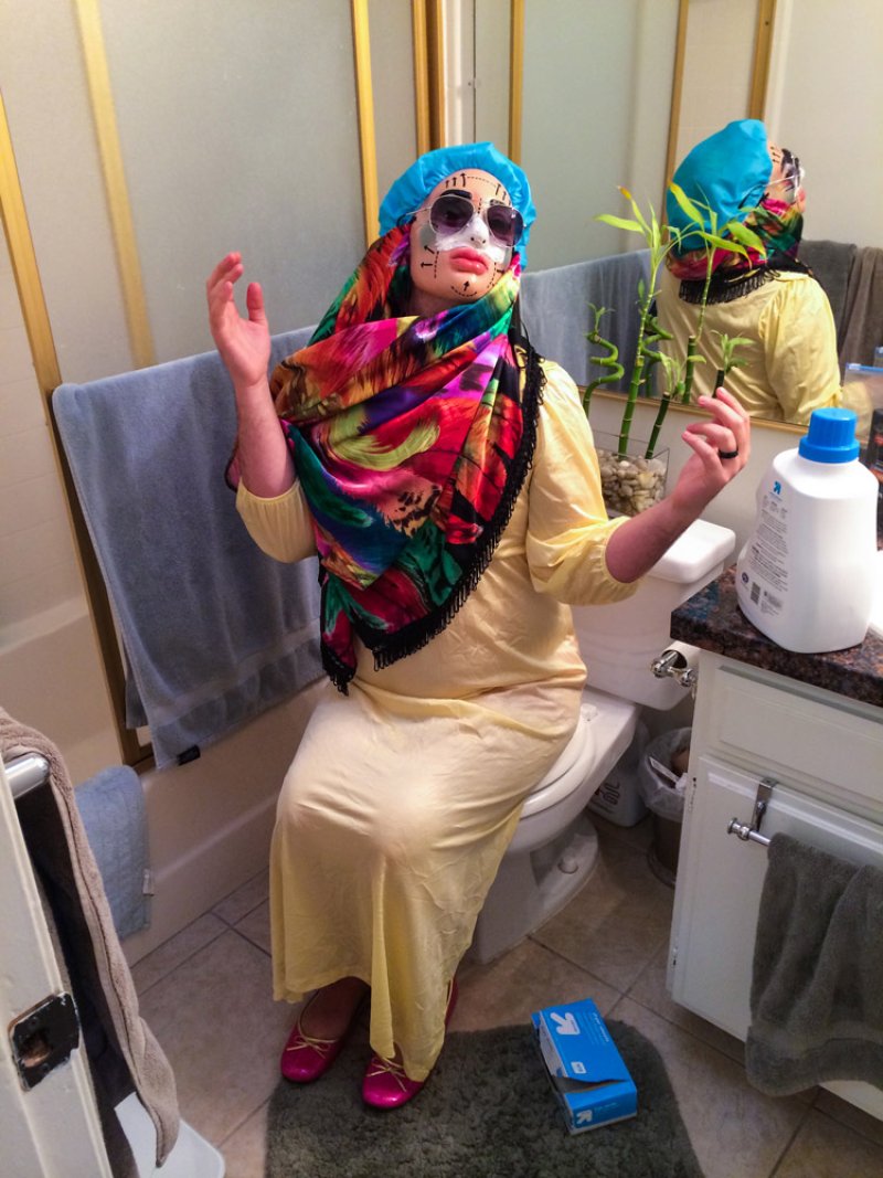 Person in yellow dress, colorful headscarf, blue shower cap, and white face mask with black markings, sitting on a toilet.