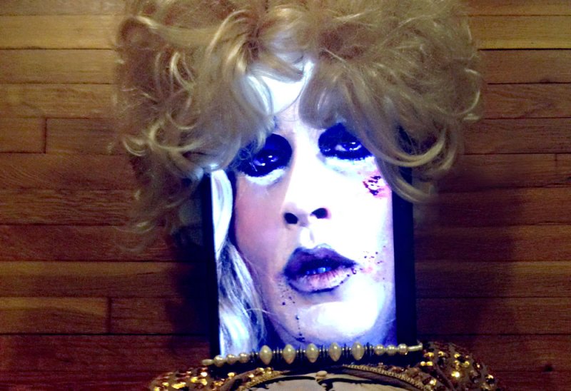 Close-up of a person with blonde hair and heavy makeup, projected onto a screen with a beaded collar.