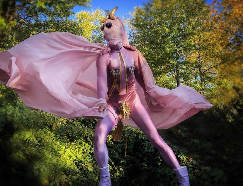Performance artist in a pink bodysuit and flowing cape, with a masked face and gold accents, against a vibrant green forest.