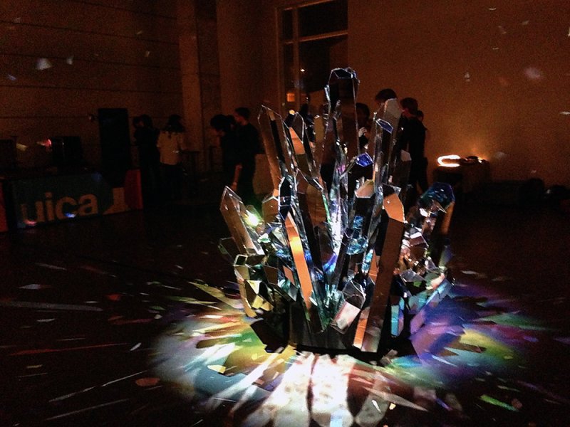 Sculpture of mirrored crystal shards reflecting colorful light onto a dark floor, with blurred figures in background.