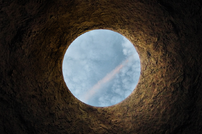 View from inside a dark, textured hole looking up at a bright blue sky with white clouds and a faint pink streak.