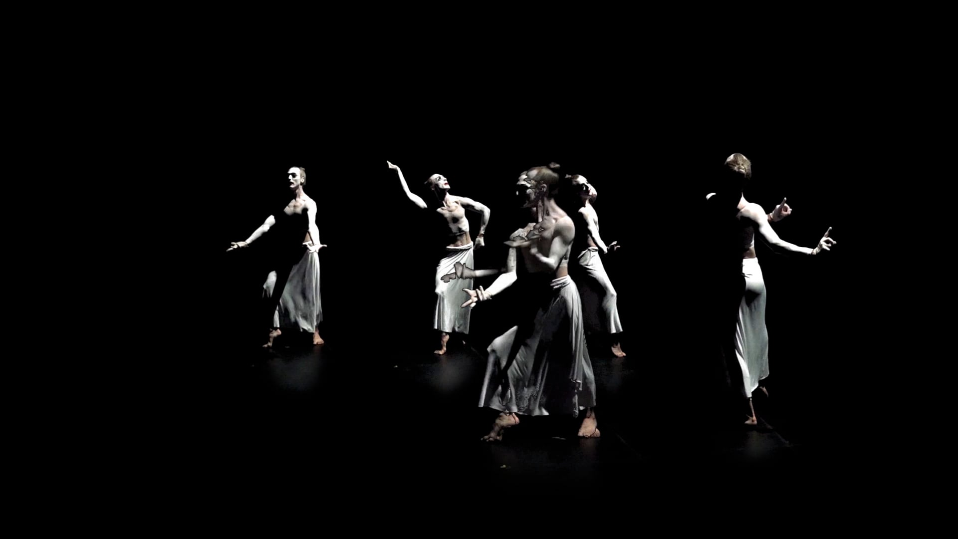 Five dancers in white skirts and painted torsos pose in a dark space, illuminated by spotlights.