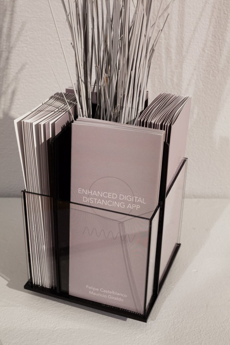 Light pink booklets with "ENHANCED DIGITAL DISTANCING APP" text in a black wire holder, with thin white reeds emerging from the top.
