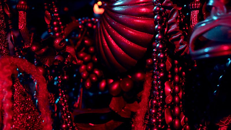 Close-up of ornate, dark red and black beaded objects, some textured and spiraled, illuminated by deep red light.