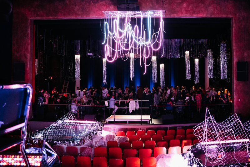 A theater stage with a large party, glowing neon chandelier, and silver streamers. Red seats and crumpled wire frames in foreground.