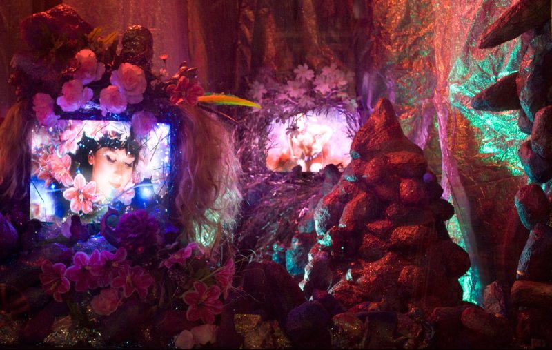 Mixed media installation with glowing screens, flowers, and rock-like forms under pink, purple, and green lighting.