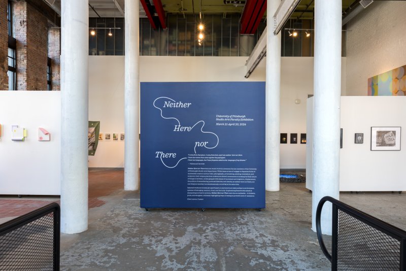 A large, dark blue panel with white text, "Neither Here nor There," stands in a gallery space with white columns.