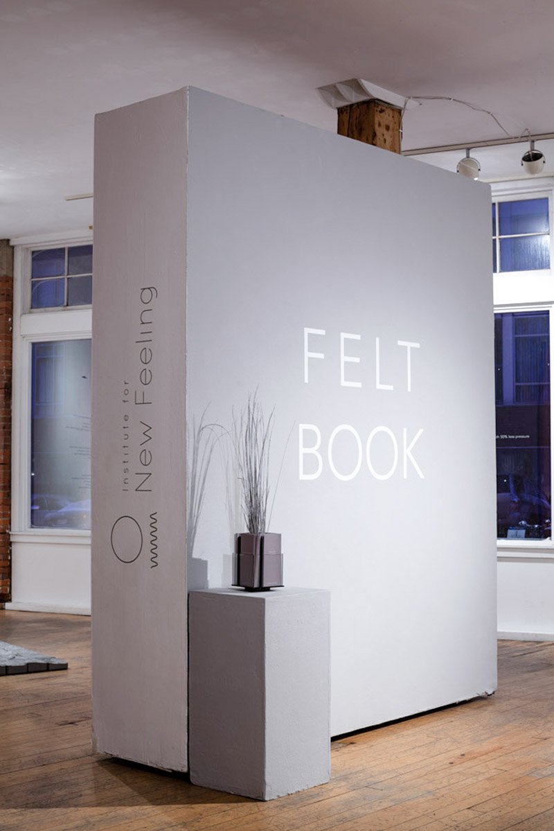 Large white book sculpture with "FELT BOOK" on the front, "Institute for New Feeling" on the spine, and a plant on a pedestal.