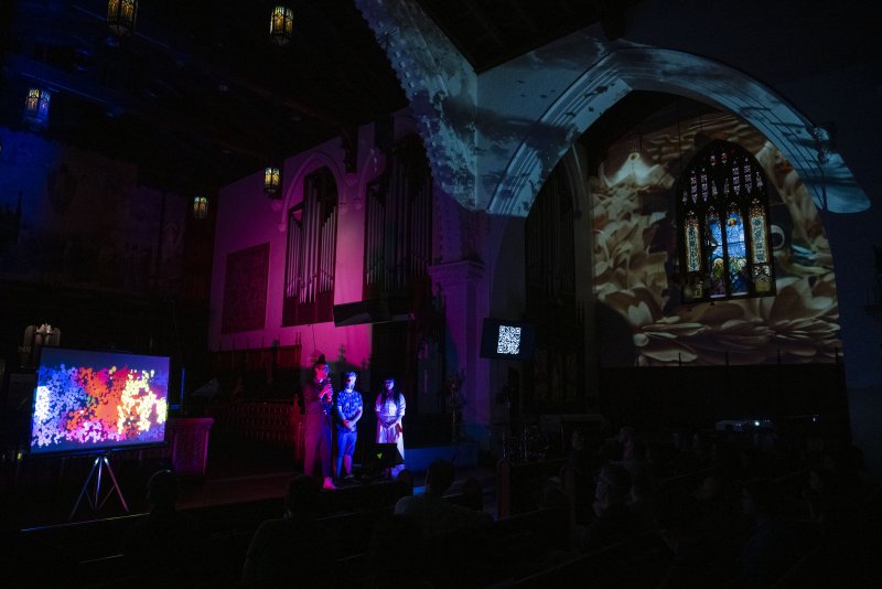 Dark church interior with three figures on stage under pink light, a colorful abstract video on a screen, and a projected image on a stained-glass window.