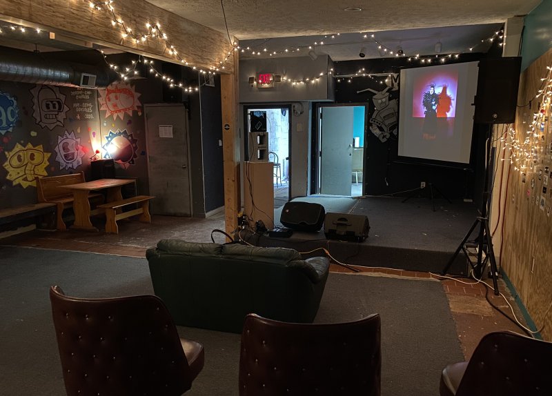 Dark room with string lights, a colorful mural, and a video projected onto a screen above a small stage.