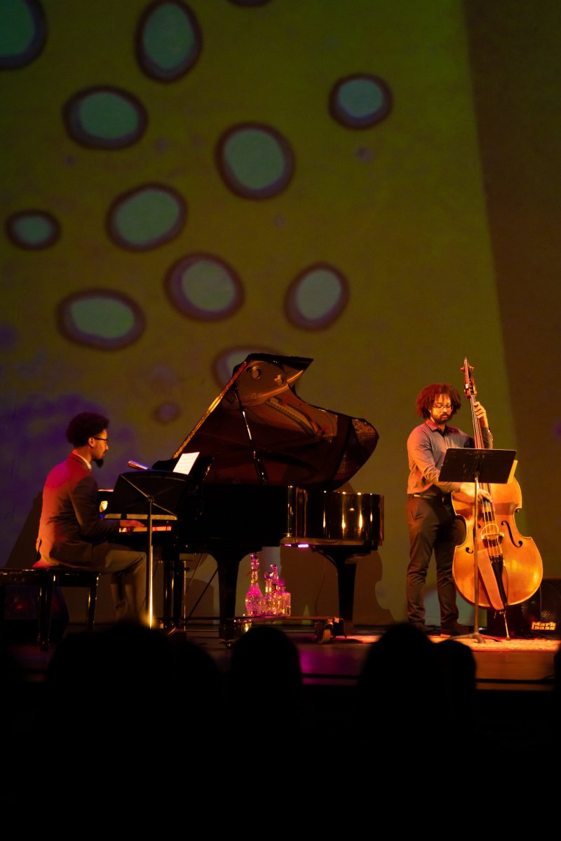Two musicians, one on grand piano and one on upright bass, perform on stage with a yellow and purple patterned backdrop.