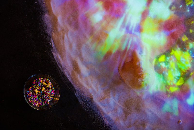 Overhead view of a bowl of colorful sprinkles next to a pile of sand illuminated by swirling pink, green, and yellow light.