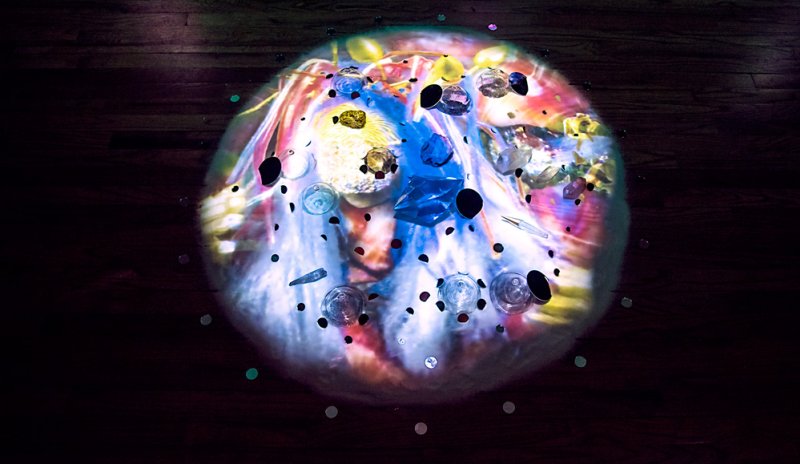 Circular projection of colorful abstract shapes, glass objects, and dark flecks on a wooden floor.